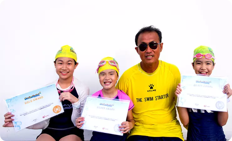 The Swim Starter students are taking a photo with the Swimming coach while holding their Swim Safer Stage 1 and SwimSafer Stage 2 Certificates